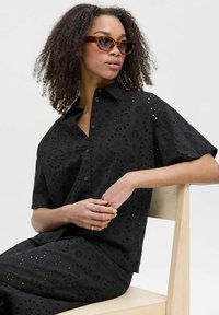 Camisa negra bordada con mangas cortas y cierre de botón, con detalles de recortes florales. Acompañada de gafas de sol de carey.