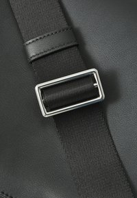 Sangle en matériau synthétique noir avec une boucle rectangulaire en métal argenté et une surface texturée, présentant un accent en cuir noir au centre.