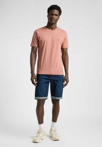 Jasnoróżowa koszulka z krótkim rękawem, granatowe szorty dżinsowe i beżowe trampki. Zawiera małe logo na piersi, luźny krój.