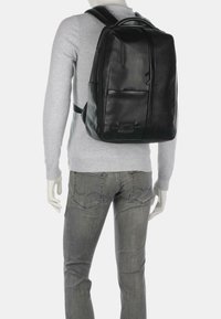 Zaino in pelle nera con un design strutturato, dotato di molteplici tasche con zip e spallacci regolabili, esposto su una figura.