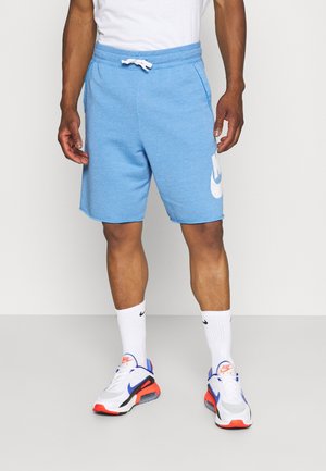 Mężczyzna w jasnoniebieskich szortach Nike z białym sznurkiem, białych skarpetkach Nike oraz białych, niebiesko-czerwonych butach Nike Air Max stojący na szarej podłodze.