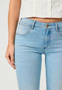 Jean en denim bleu clair au design ajusté, légères marbrures sur les cuisses, cinq poches et fermeture à bouton en cuivre.