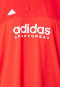 Červené sportovní tričko Adidas s bílým textovým logem, vertikálně pružnou texturou a malým bílým logem na levém rameni.