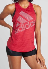 Červený sportovní top bez rukávů s černými ramínky, který má velké bílé pruhované logo Adidas. Materiál vypadá lehce a má hladkou texturu.