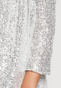 Zbliżenie na przedramię noszące białą odzież gęsto pokrytą małymi, błyszczącymi srebrnymi cekinami odbijającymi światło.