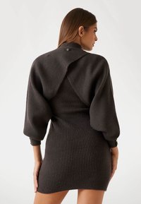 Robe en tricot côtelé noir avec une silhouette ajustée, des manches ballon et un dos ouvert. Présente des accents en métal doré au niveau des poignets.