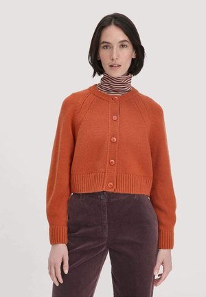Korte oranje gebreide cardigan met een ribbelboord aan de onderkant en aan de mouwen, voorzien van vijf knopen aan de voorkant. Gecombineerd met een gestreepte coltrui en donkere broek.