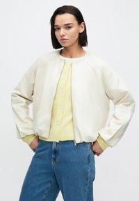 Biela bomber bunda z umelej kože s rebrovanými manžetami a zipsom, v kombinácii so žltým kardigánom a modrými džínsami.