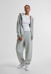 Lys grå hettegenser og matchende joggebukser laget av mykt stoff. Hvit tanktop under. Hvit joggesko med snøring.