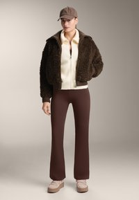 Femme portant une casquette marron, une veste polaire marron foncé, un pull à fermeture éclair crème, un pantalon marron et des chaussures compensées beige, posant devant un fond neutre.