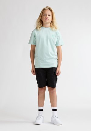 Tricou verde mentă cu mâneci scurte, asortat cu pantaloni scurți cargo negri. Adidași albi și șosete cu dungi alb-negru completează ținuta.