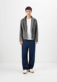 Grå ulljakke med store lommer, lagdelt over en hvit skjorte. Kombinert med løstsittende blå jeans og beige joggesko. Minimalistisk design.
