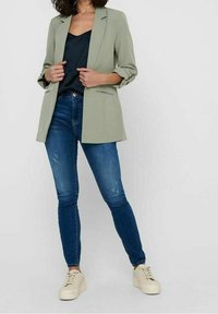 Frau in einem salbeigrünen Blazer über einem schwarzen Oberteil, mit verwaschenen blauen Skinny Jeans und beigen Plateausneakern, posiert vor einem schlichten Hintergrund.