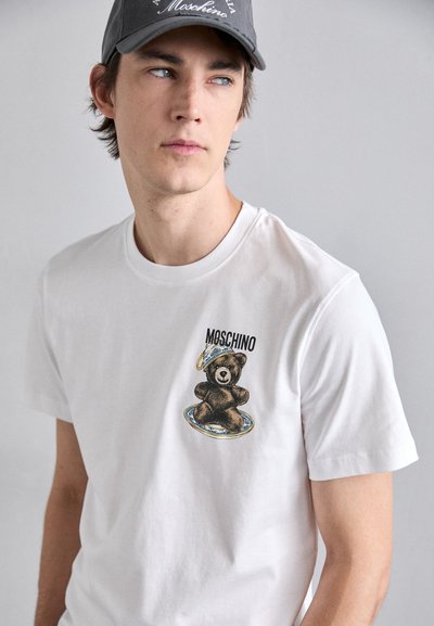 Balts kokvilnas T-krekls ar lāča grafiku brūnās un melnās krāsās, ar uzrakstu "Moschino". Īsām piedurknēm un apaļu kakla izgriezumu.