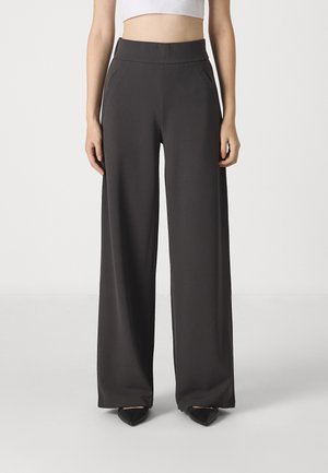 Kvinde iført sorte bukser med høj talje og vide ben, kombineret med en hvid cropped top og sorte sko med spids tå.