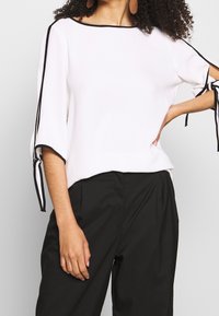 Blouse blanche avec des bordures noires, manches larges et liens aux poignets, assortie à un pantalon noir. Texture lisse et design décontracté.