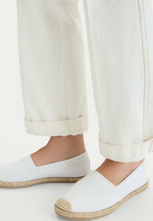 Füße in weißen Canvas-Schuhen zum Hineinschlüpfen mit geflochtenem Jutebesatz, kombiniert mit cremefarbenen, umgeschlagenen Hosen vor einem einfarbigen Hintergrund.