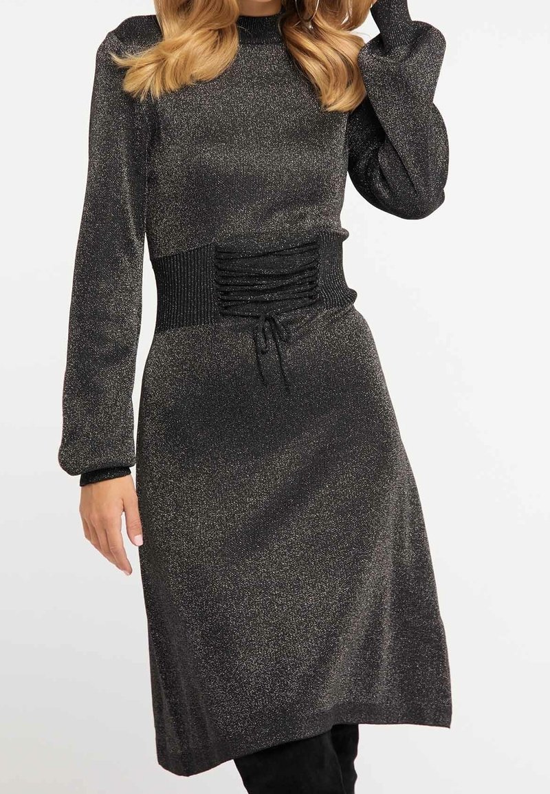 Femme portant une robe noire pailletée à manches longues et longueur genou, avec une ceinture froncée large et un détail à nouer à l'avant.