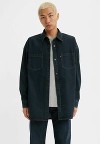 Camisa chaqueta de mezclilla oscura con cierre de botones, dos bolsillos frontales y un cuello estructurado, superpuesta a una camiseta gris clara.