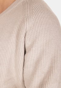 Pullover di maglia beige con texture a coste, scollo a V e spalle leggermente cadenti. Il tessuto appare morbido e caldo.
