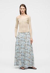 Γυναίκα στέκεται φορώντας μπεζ ριμπ πλεκτή ζακέτα με κουμπιά και μακριά μπλε φούστα με σχέδια, με μαύρα σανδάλια, με τα χέρια στις τσέπες.