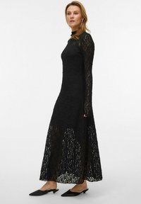 Vestido largo maxi de encaje negro con mangas largas, escote alto y textura transparente. Combinado con mules negros de punta afilada, sobre un fondo liso.