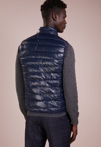 Gilet imbottito blu navy con finitura lucida, texture trapuntata e colletto alto, indossato sopra un maglione grigio, vista posteriore.