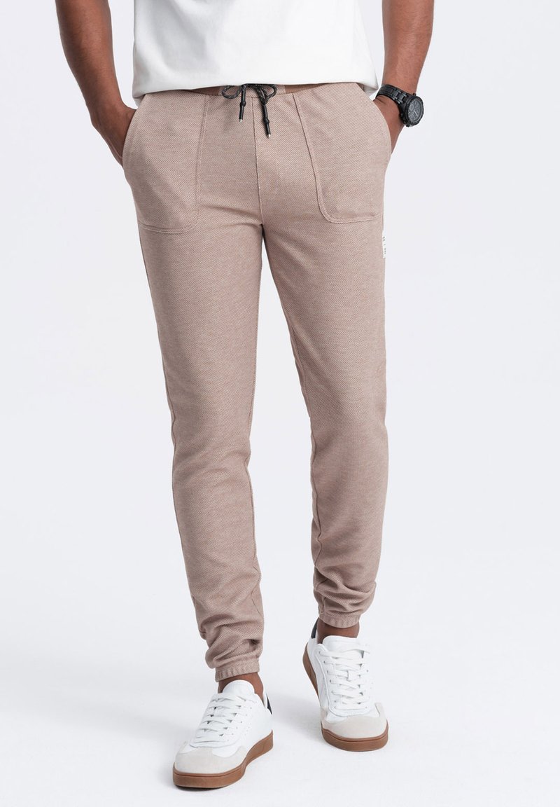Jasnobrązowe joggery z teksturowanej tkaniny, elastycznym pasem i regulowanym sznurkiem. Posiadają kieszenie boczne i zwężane nogawki.