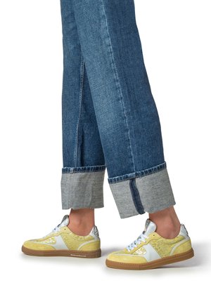 Gele suède sneakers met zilveren accenten en witte veters, gecombineerd met blauw spijkerbroek met omgeslagen boorden en een donkerder zoom.