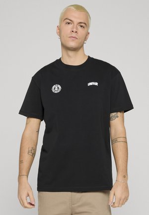 Junger Mann mit kurzen blonden Haaren, trägt ein schwarzes "UNFAIR"-T-Shirt und beige Hosen, zeigt Tätowierungen auf beiden Armen vor neutralem Hintergrund.