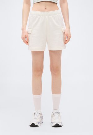 Oseba, ki nosi bež cargo kratke hlače z elastičnim pasom, svetlo roza nogavice z okroglim izrezom, bele in bež superge, stoji ob enobarvnem belem ozadju.