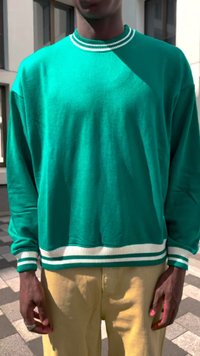 Personne portant un pull-over vert à manches longues avec des rayures blanches au col, aux poignets et à l'ourlet, associé à un pantalon jaune, se tenant à l'extérieur.
