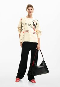 Crèmekleurige trui met cartoonontwerp, zwarte broek en roze schoenen. Zwarte handtas met textuurpatroon en rode accenten. Casual outfit.