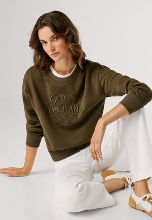 Donna con capelli castani ondulati seduta a terra che indossa una felpa verde oliva "Pepe Jeans", pantaloni bianchi e sneakers beige e bianche su sfondo neutro.