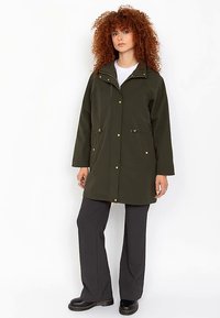 Cappotto impermeabile verde scuro con collo alto, due tasche frontali e bottoni a pressione dorati. Abbinato a pantaloni grigi a gamba larga e stivali neri.