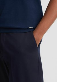 Marinblå bomulds Sweater med ribbet kant, der har et lille sølvlogo tag på siden. Kombineret med mørke bukser, der fremhæver kontrasten i teksturen.
