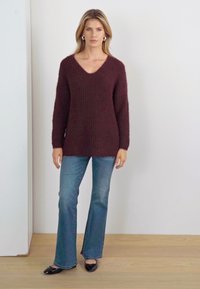 Burgunderfarbener, übergroßer Strickpullover mit V-Ausschnitt und langen Ärmeln, kombiniert mit hellblauen, ausgestellten Jeans und schwarzen Ballerinas. Hintergrund aus Holzfußboden.