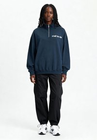 Sweat-shirt bleu marine zippé avec le texte « c'est la vie », associé à un pantalon cargo noir et des baskets blanches. Tissu doux, coupe décontractée, poignets côtelés.