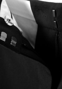 Nærbilde av svart plaggliv med to krooks- og barlås, svart glidelås og en hvit knapp på innsiden av foret.