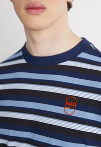 Marinblå, svart och ljusblå randig t-shirt med ribbad krage, prydd med en orange broderad logotyp på bröstet, tillverkad av mjukt bomullsmaterial.