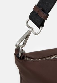 Détail d'un sac en cuir marron présentant un fermoir et une fermeture éclair en métal argenté, avec une sangle texturée noire et des accents en cuir marron.