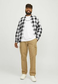 Camisa de franela a cuadros en blanco y negro sobre una camiseta blanca, combinada con pantalones cargo beige y zapatillas blancas sobre un fondo neutro.