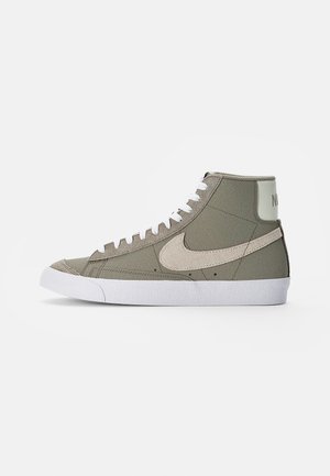 Baskets montantes vert olive avec semelle et lacets blancs, arborant un swoosh Nike beige texturé sur le côté.