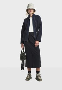 Chaqueta de forro polar azul marino con cuello alto, combinada con una camisa blanca y una falda negra. Zapatillas verde oliva y un pequeño bolso verde completan el look.
