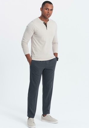 Beige overhemd met lange mouwen en henley-kragen met zwarte knoopaccenten, gecombineerd met marineblauwe broek en lichtbruine sneakers. Staande houding met handen in de zakken.