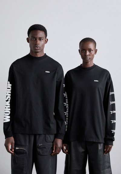 Zwei Personen tragen schwarze Langarmshirts mit weißen Druckmotiven. Auf dem einen Shirt steht "WORKSHOP" auf dem Ärmel; das andere hat verschiedene kleine Grafiken.