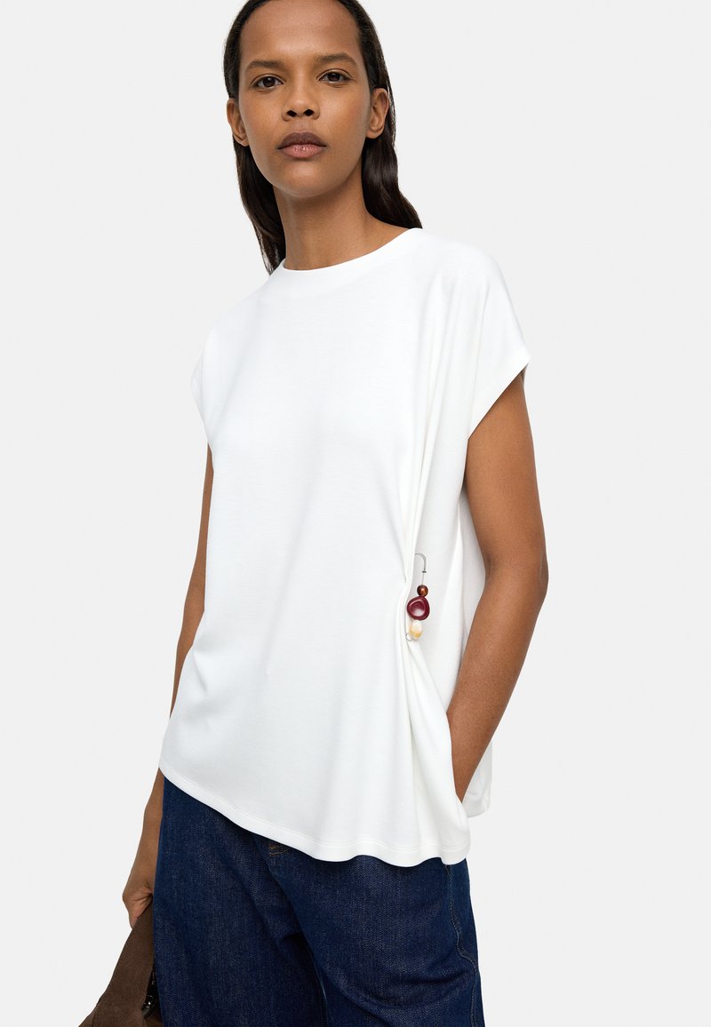 Biała bluzka z krótkim rękawem o luźnym kroju, z boczną kieszonką ozdobioną kolorowymi koralikami, zestawiona z niebieskimi dżinsami.