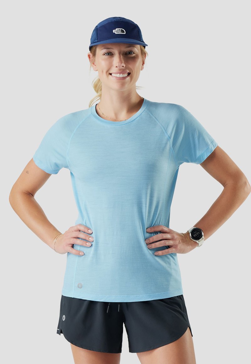 Femme souriante en T-shirt bleu clair, short noir, casquette bleue et montre connectée blanche, debout les mains sur les hanches devant un fond uni.