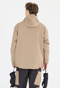 Beige vedenkestävä takki hupulla, joustavilla hihansuilla ja kiristyshelmalla. Mustat hanskat vasemmassa kädessä, ylle puettuna tummat housut, joissa on vaaleita laikkuja.