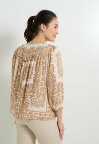 Blusa ligera beige con estampado de paisley dorado, mangas globo de ¾ y detalle de yugo fruncido en la parte posterior. Textura de tejido suave.
